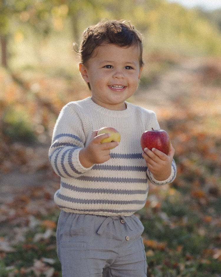 A smiling young child outdoors, dressed in the Quincy Mae rex sweater in slate stripe and gray pants, holds a red apple in one hand and a yellow apple in the other, surrounded by autumn leaves on the ground.