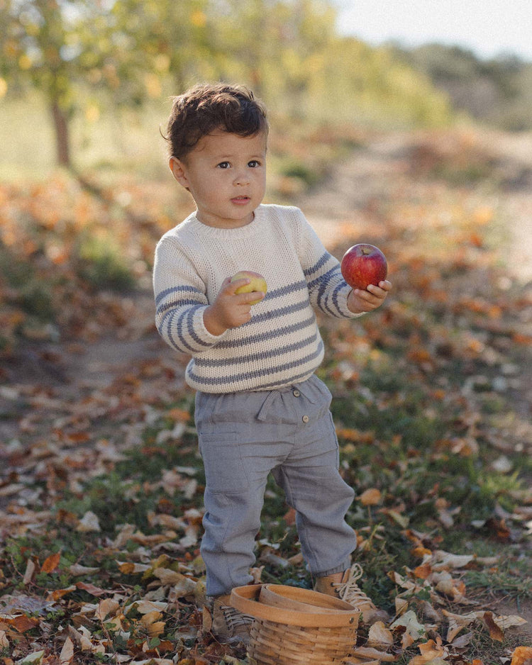 A young child stands outdoors on a leaf-covered path, wearing the quincy mae rex sweater in slate stripe. They hold a red apple in one hand and a yellow apple in the other, with a small wooden basket at their feet.