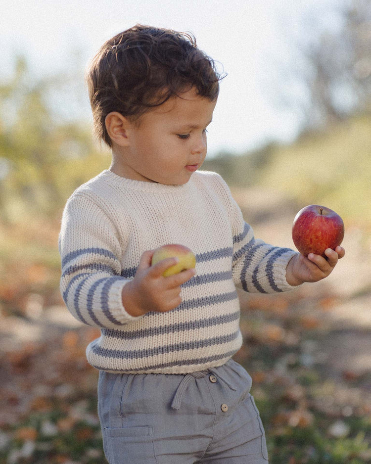 A young child with short brown hair, wearing the quincy mae rex sweater in slate stripe and gray pants, stands outdoors holding a red apple in one hand and a yellow apple in the other.