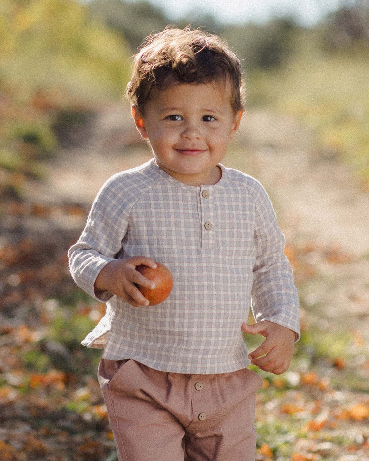 A young child stands outdoors on a sunlit path, smiling at the camera and holding an apple, wearing the quincy mae zion shirt in blue gingham and pink pants.