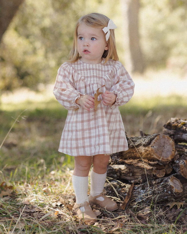 A young girl with blonde hair, wearing a pink plaid dress, a white bow, and the quincy mae socks set in natural + dusty rose, stands outdoors on grass near a stack of logs, looking up with a curious expression.