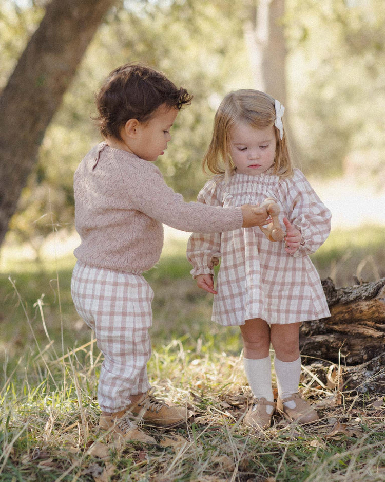 Two young children stand outdoors on grass, both dressed in matching quincy mae woven pant in rustic plaid, made from organic cotton in soft tones. One child offers a toy to the other, who gazes at it with curiosity. Trees and sunlight fill the background, highlighting the relaxed fit of their pants.