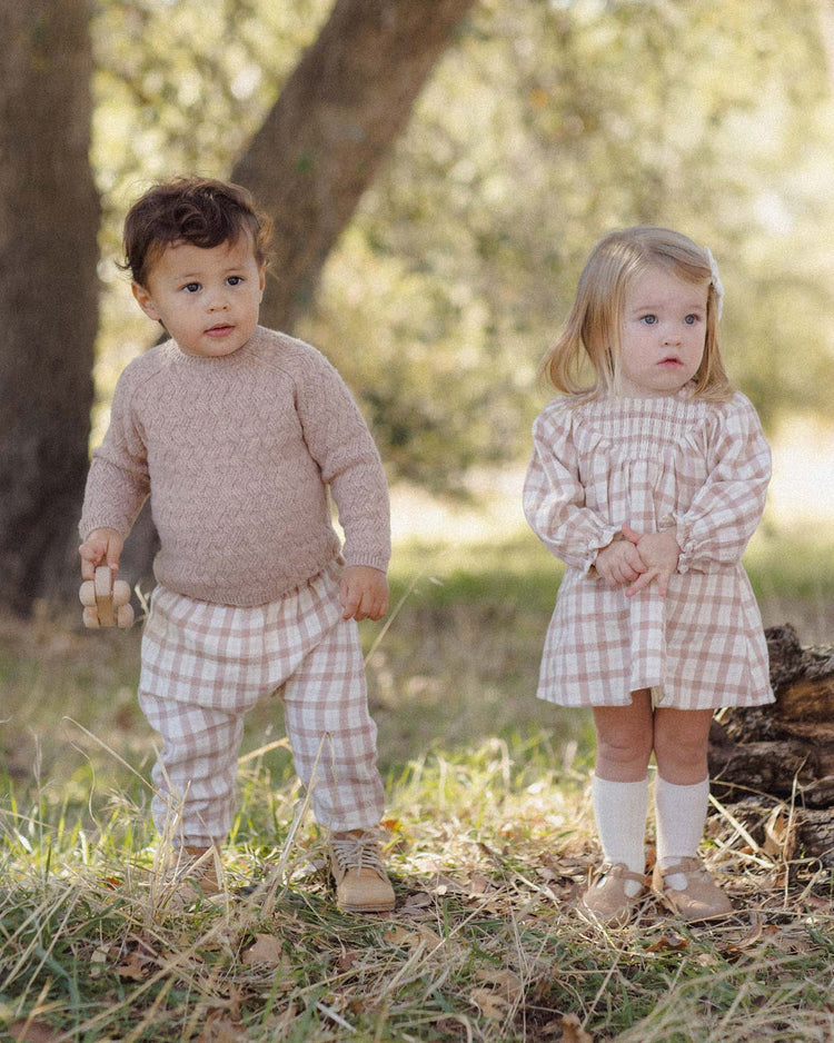 Two young children stand outdoors in a grassy, wooded area. Both are wearing coordinating beige and white "woven pant in rustic plaid" outfits from Quincy Mae, crafted from organic cotton and paired with neutral shoes. One child holds a small wooden toy while the other clasps their hands in front.