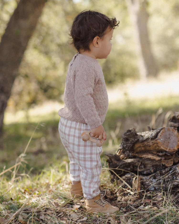 A young child wearing the quincy mae woven pant in rustic plaid stands outdoors on grass near cut tree logs, dressed in a cozy knit sweater and boots, holding a wooden toy while looking to the side with trees softly blurred in the background.