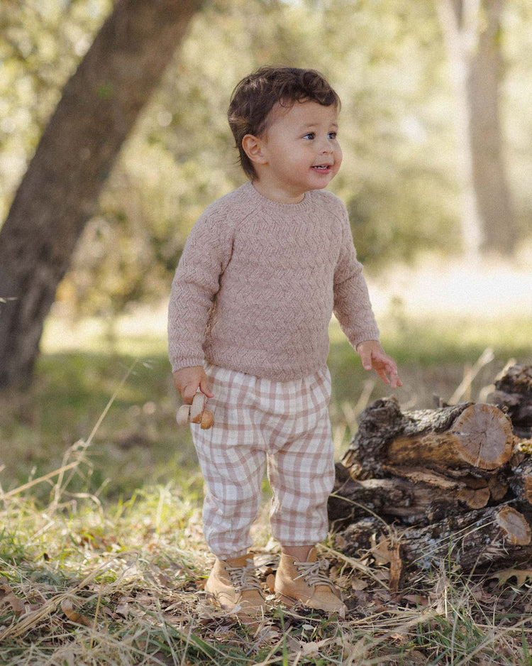 A young child stands outdoors on the grass near a pile of logs, smiling. The child wears a light brown organic cotton sweater, tan boots, and the quincy mae woven pant in rustic plaid, holding a small object. Sunlight filters through the trees in the background.