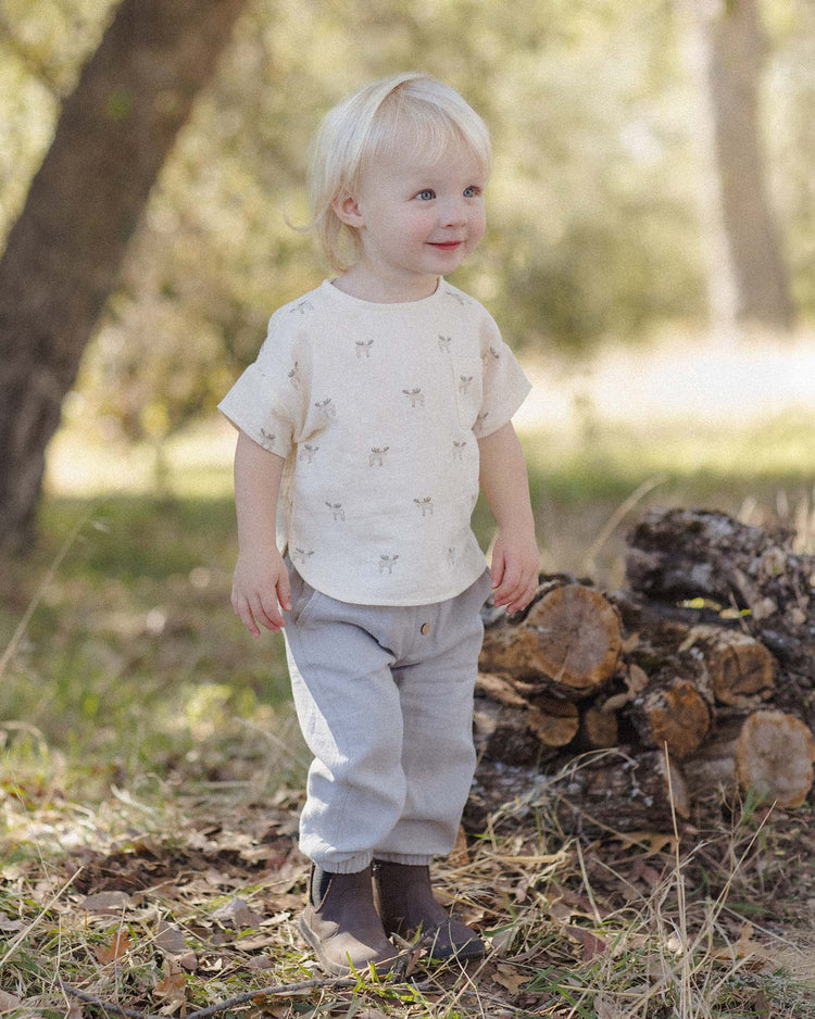 A young child with blonde hair stands outdoors on grass near a small stack of cut logs, smiling. The child wears a light-colored shirt, the quincy mae giovani pant in cloud—a woven gauze pant with an elastic waistband—and brown boots, while sunlight filters through trees in the background.
