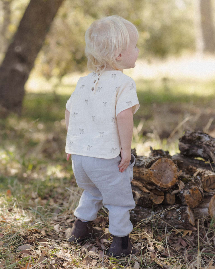 A toddler with blond hair, wearing a cream shirt and the quincy mae giovani pant in cloud—crafted from organic cotton with an elastic waistband—stands outdoors on grass near a stack of cut wood, facing away. The wooded scene is sunlit.