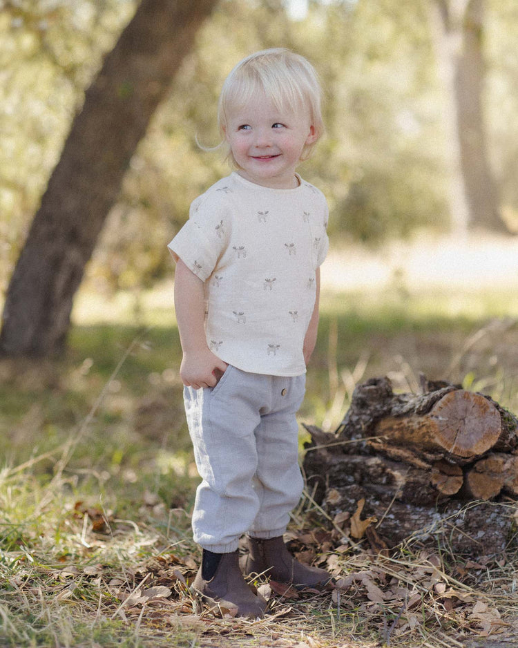 A young child with blonde hair stands outdoors near a pile of logs, wearing a light-colored shirt, quincy mae’s giovani pant in cloud—gray woven gauze pants with an elastic waistband—and brown boots. The background shows trees and grass in soft sunlight.