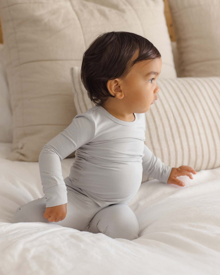 A young child in the quincy mae bamboo pajama set in cloud sits on a white bed with large beige pillows, looking to the side with a curious expression.