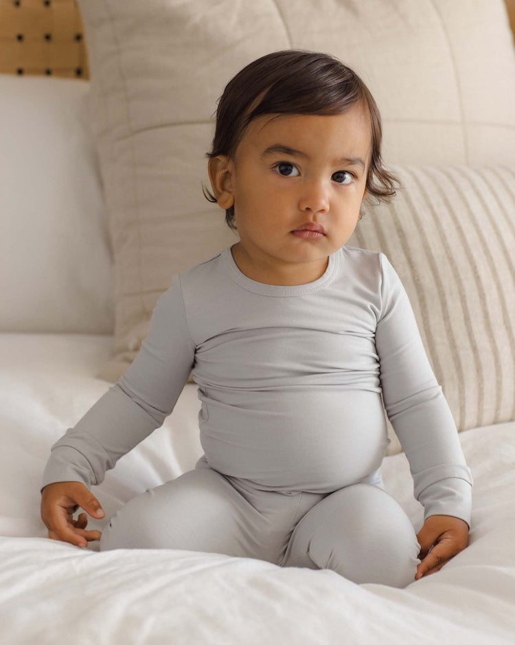 A young child with dark hair sits on a bed, wearing the quincy mae bamboo pajama set in cloud. The background features neutral-colored pillows and bedding. The child gazes slightly to the side with a calm expression.