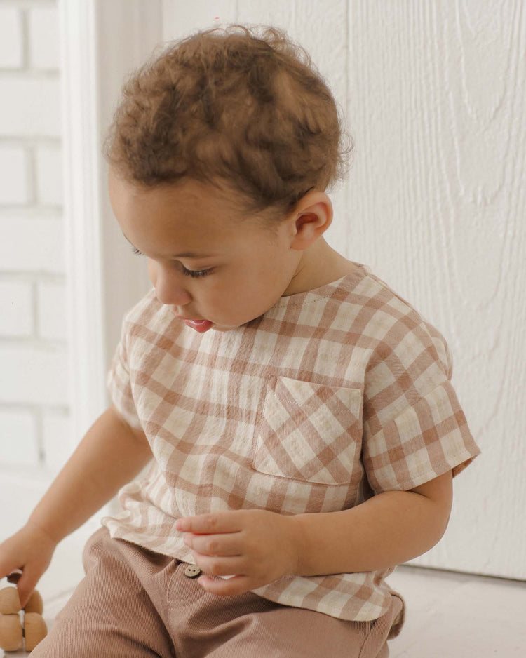 A young child with curly hair sits indoors, looking down while holding a toy, dressed in the quincy mae woven boxy top in rustic plaid (beige and white) paired with light brown pants.