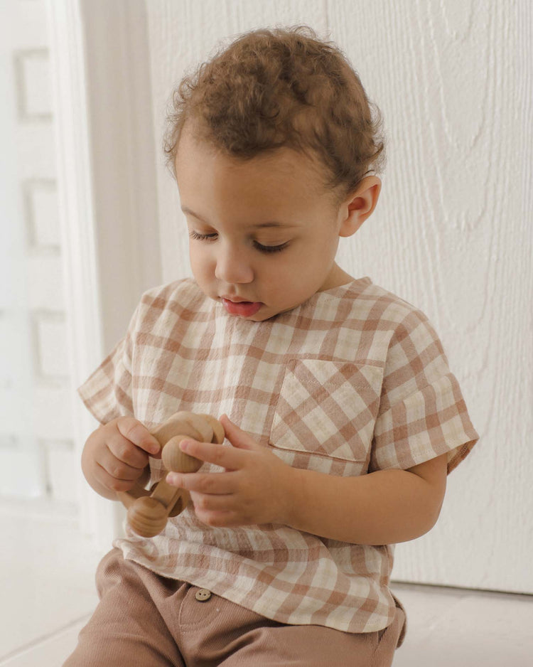 A young child wears the quincy mae woven boxy top in rustic plaid, made from organic cotton, while sitting in front of a white textured wall and holding a small wooden toy.