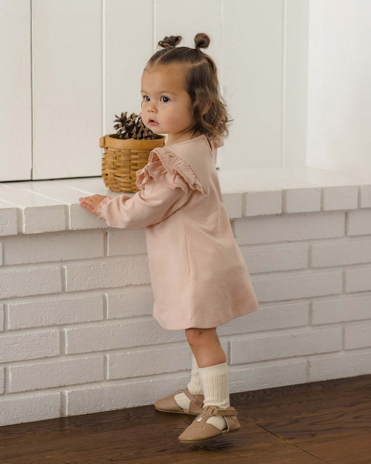 A toddler with short brown hair in a pink dress, beige shoes, and the Quincy Mae socks set in natural + dusty rose stands beside a white brick fireplace, holding the edge. A small woven basket with pinecones rests on the ledge.