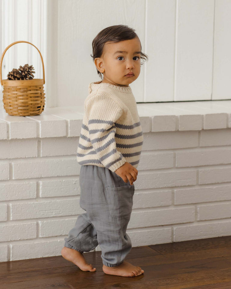 A young child with dark hair, wearing a striped sweater and the quincy mae luca pant in slate—comfortable woven pants with an elastic waistband—stands barefoot on a wooden floor near a white brick wall. A small basket with pine cones sits on the mantle behind them.