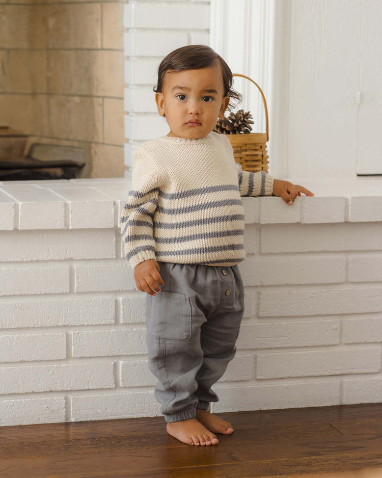 A toddler with dark hair stands barefoot on a wooden floor, resting one hand on a white brick fireplace, wearing a cream and gray striped sweater and the quincy mae luca pant in slate with an elastic waistband.
