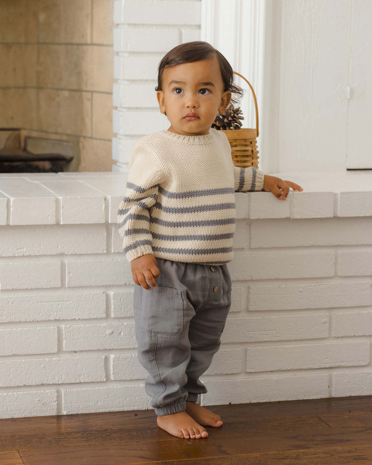 A toddler with short brown hair stands barefoot on a wooden floor in front of a white brick fireplace, looking slightly to the left. They are wearing comfortable luca pants in slate by quincy mae, paired with a cream and gray striped sweater featuring an elastic waistband.