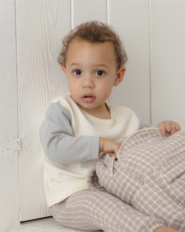A young child with curly hair sits against a white wall, looking at the camera while holding a plaid-patterned bag. The child wears the quincy mae long sleeve raglan tee in cloud, made from organic cotton, along with relaxed fit plaid pants.