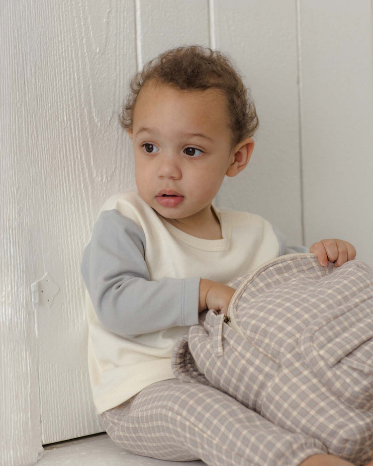 A young child with short curly hair sits on the floor beside a white wall, looking to the side and holding a plaid-patterned backpack. They wear the quincy mae long sleeve raglan tee in cloud, crafted from soft organic cotton for comfortable, casual style.