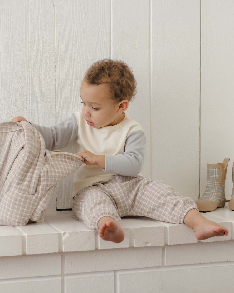 A toddler in a quincy mae woven pant in blue gingham and a cream top sits on a white ledge, looking inside a plaid backpack. Beige plaid boots rest beside them against a white wood wall.
