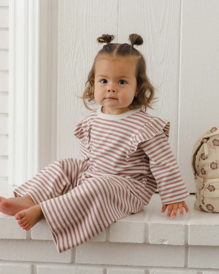 A toddler with brown hair in two buns sits barefoot on a white surface, dressed in the quincy mae ruffle cozy set in mulberry stripe made from organic cotton pajamas. A patterned bag is placed beside them.
