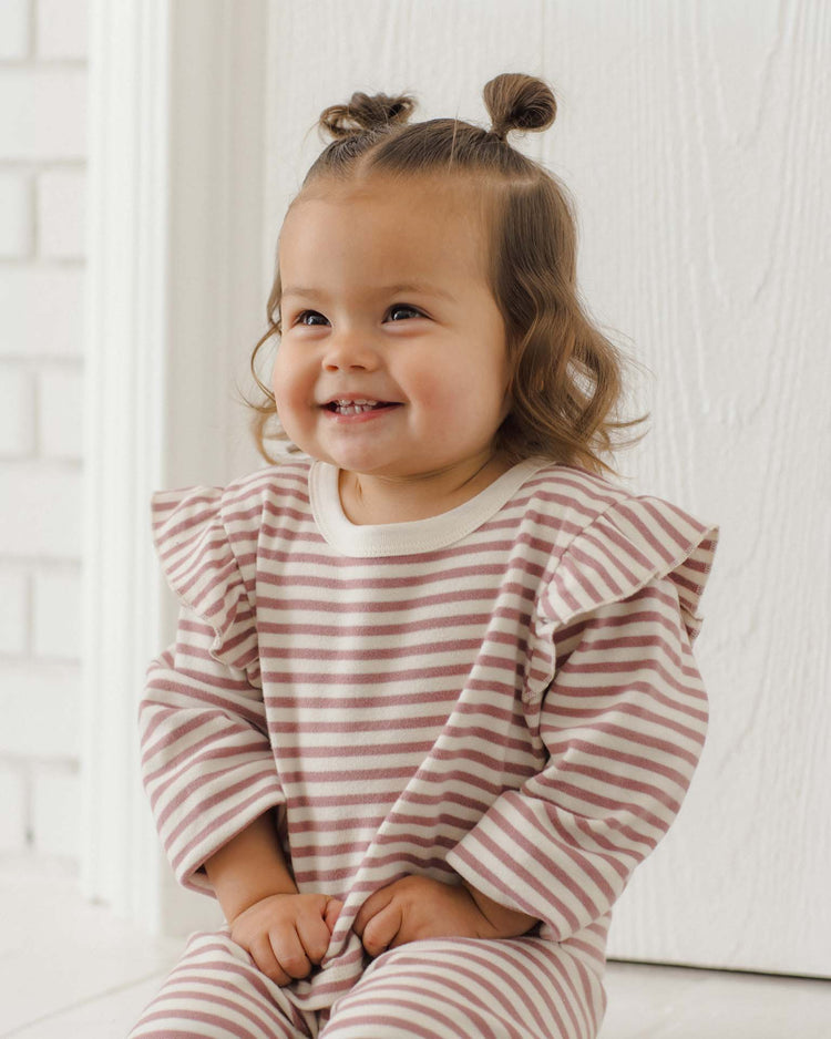 A smiling toddler with brown hair styled in two small buns wears the quincy mae ruffle cozy set in mulberry stripe, crafted from soft organic cotton, while sitting indoors against a light-colored wall and door.
