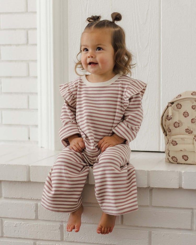 A toddler with brown hair in two buns sits barefoot on a white brick ledge, wearing the quincy mae ruffle cozy set in mulberry stripe, next to a beige backpack decorated with flower prints.