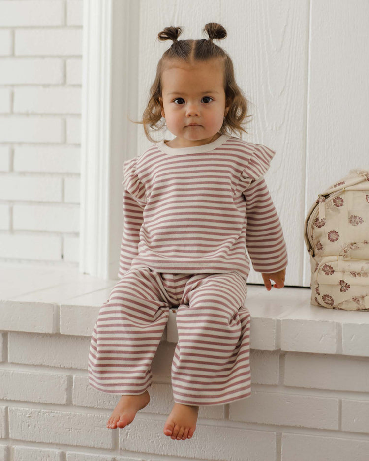 A toddler with light brown hair in two buns sits barefoot on a white brick ledge, wearing the quincy mae ruffle cozy set in mulberry stripe. A floral bag sits beside them.