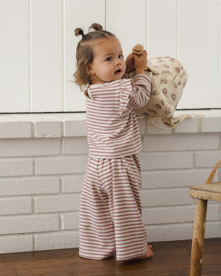 A toddler wearing the quincy mae ruffle cozy set in mulberry stripe stands by a white brick wall, holding a patterned bag and looking over their shoulder.