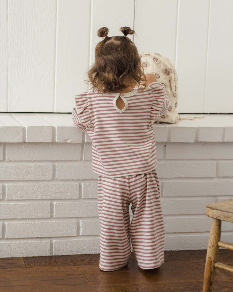 A young child with brown hair in pigtails, dressed in the Quincy Mae ruffle cozy set in mulberry stripe, stands facing a white brick wall and reaches toward a patterned bag on the ledge.