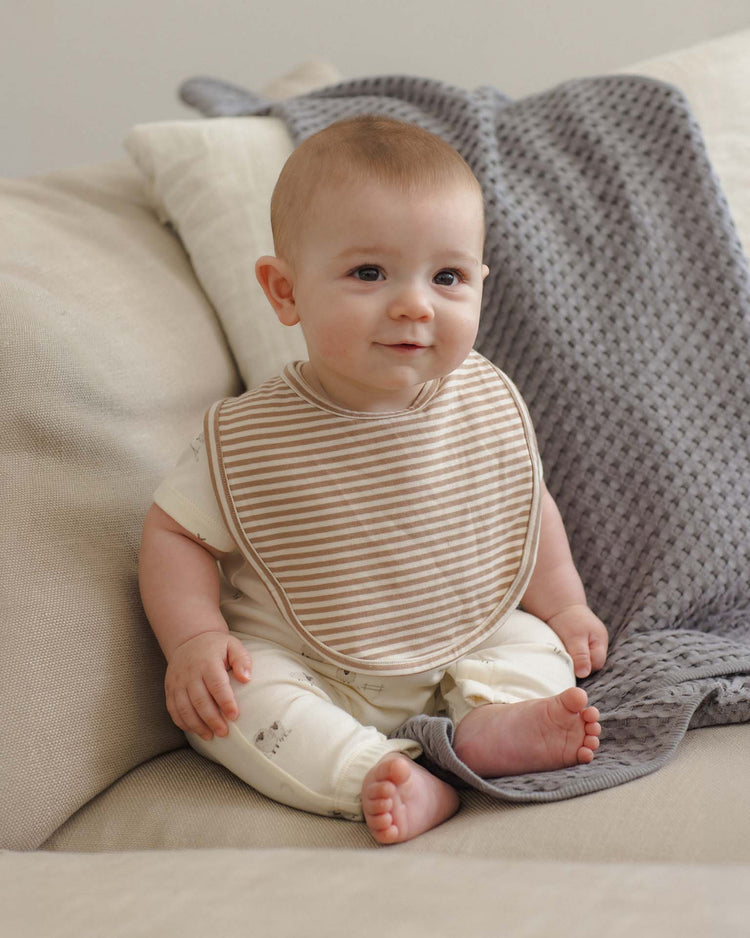 A baby wearing the quincy mae jersey snap bibs in bunnies + almond stripe and light-colored clothes sits on a beige couch next to a textured gray blanket, smiling softly.