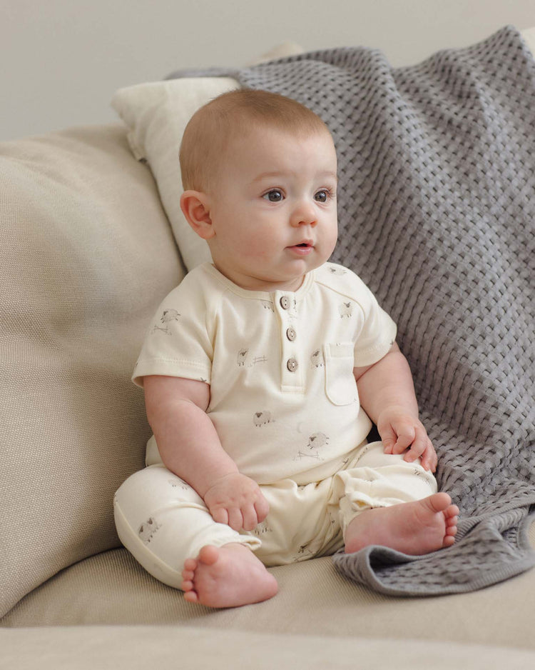 A baby wearing the quincy mae short sleeve jumpsuit in sheep sits on a beige couch next to a textured gray blanket, looking slightly to the right with a curious expression.