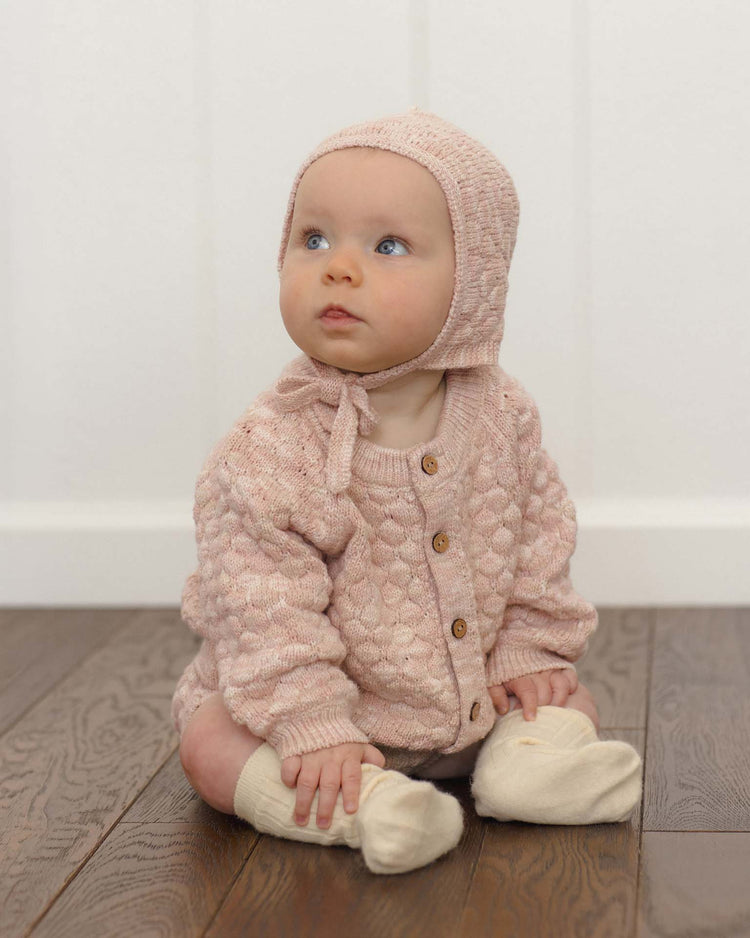 A baby wearing a quincy mae bubble knit cardigan in dusty rose speckle and a matching pink organic cotton bonnet sits on a wooden floor, looking up. The baby is also wearing cream-colored socks, with a white paneled wall in the background.