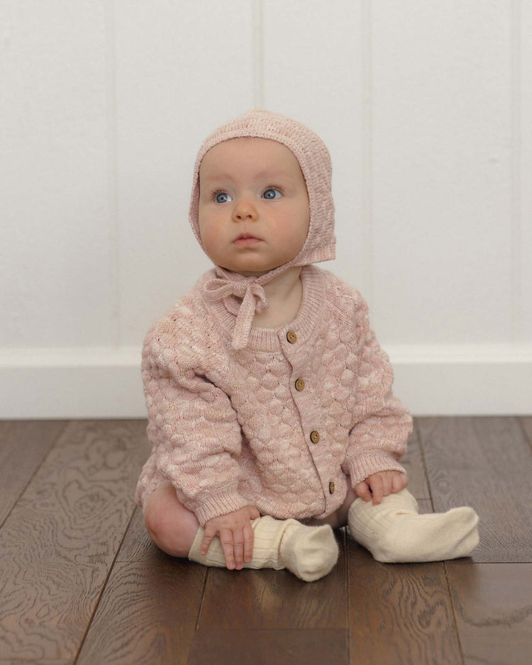 A baby wearing a pink knit bonnet, a matching cardigan, and cream socks from quincy mae sits on a wooden floor, gazing upward with curiosity. The look is finished with quincy mae’s knit bloomer in dusty rose speckle and cozy organic cotton touches for a sweet, coordinated outfit.