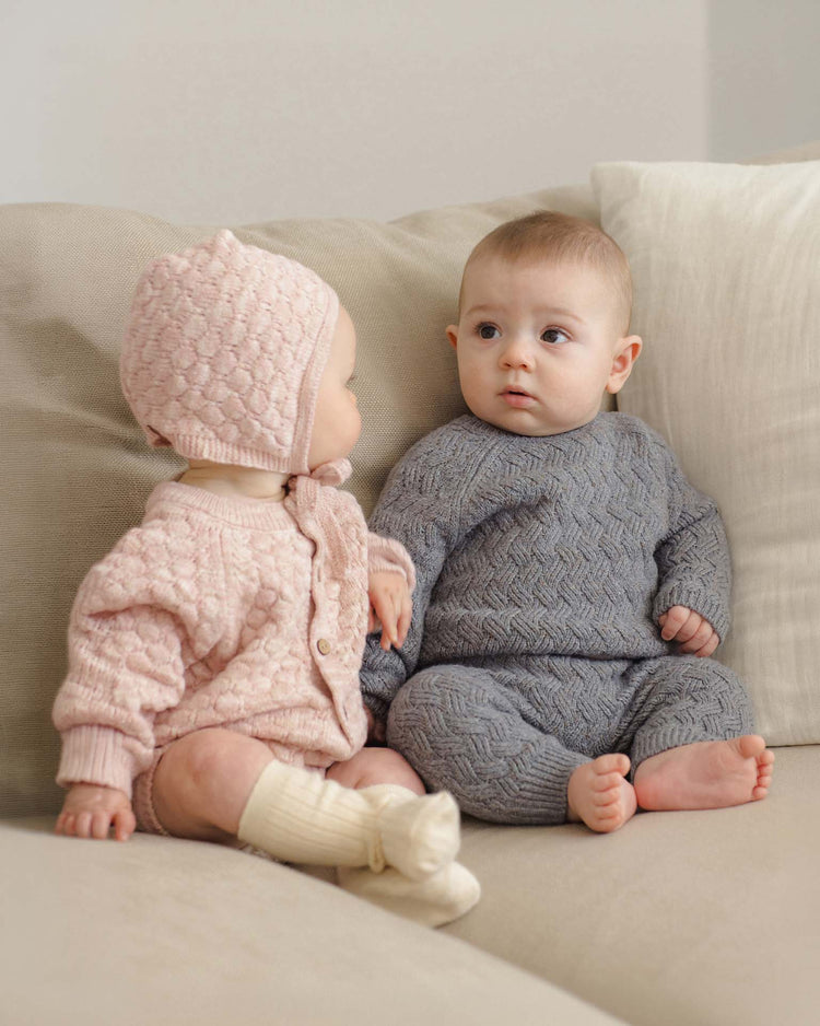 Two babies sit on a beige couch. One wears the quincy mae knit bloomer in dusty rose speckle—a pink outfit with bubble knit bloomers, matching bonnet and socks, all crafted from soft organic cotton. The other wears a gray knit sweater and pants. They face each other, appearing to interact.