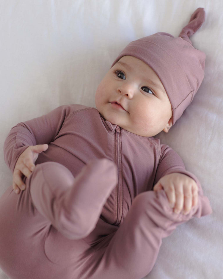 A baby wearing the quincy mae bamboo knotted baby hat in mulberry lies on a white blanket, looking up with a slight smile while holding its foot.