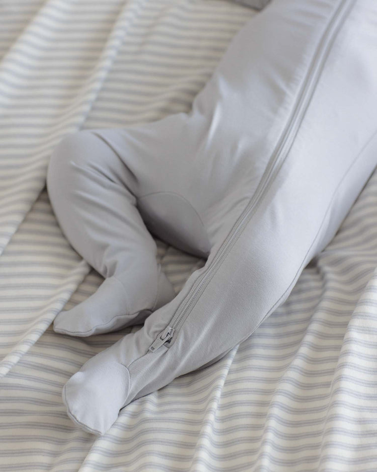 A baby wearing the Quincy Mae bamboo zip footie in Cloud lies on a bed covered with a striped sheet, with only the legs and lower torso visible.