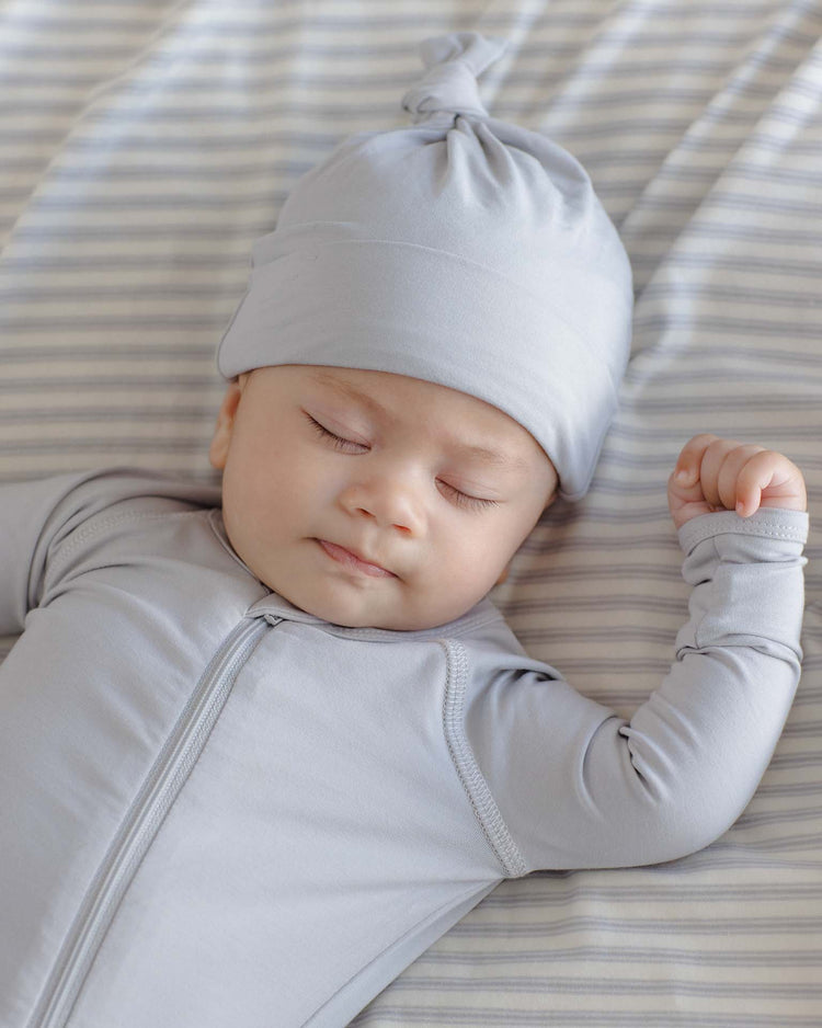 A baby in the quincy mae bamboo zip footie in cloud and a light gray hat sleeps peacefully on a striped bed sheet, with one arm raised and eyes closed.
