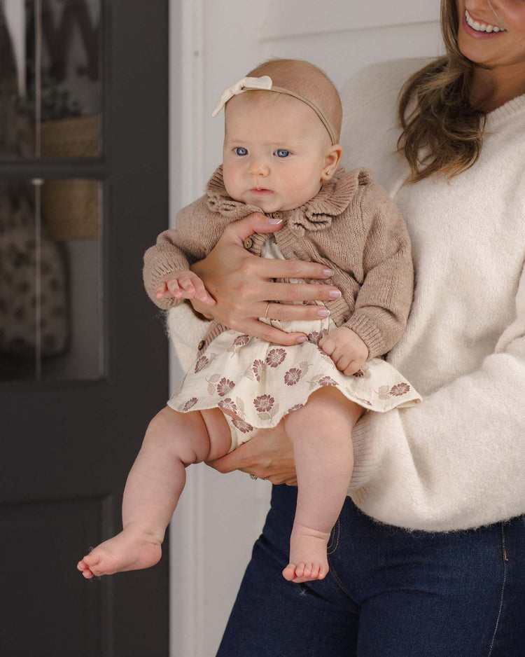 A woman in a white sweater holds a baby, who is wearing the quincy mae ruffle collar cardigan in heathered almond—crafted from recycled polyester—along with a floral dress and a matching headband. The baby looks forward while being held securely.