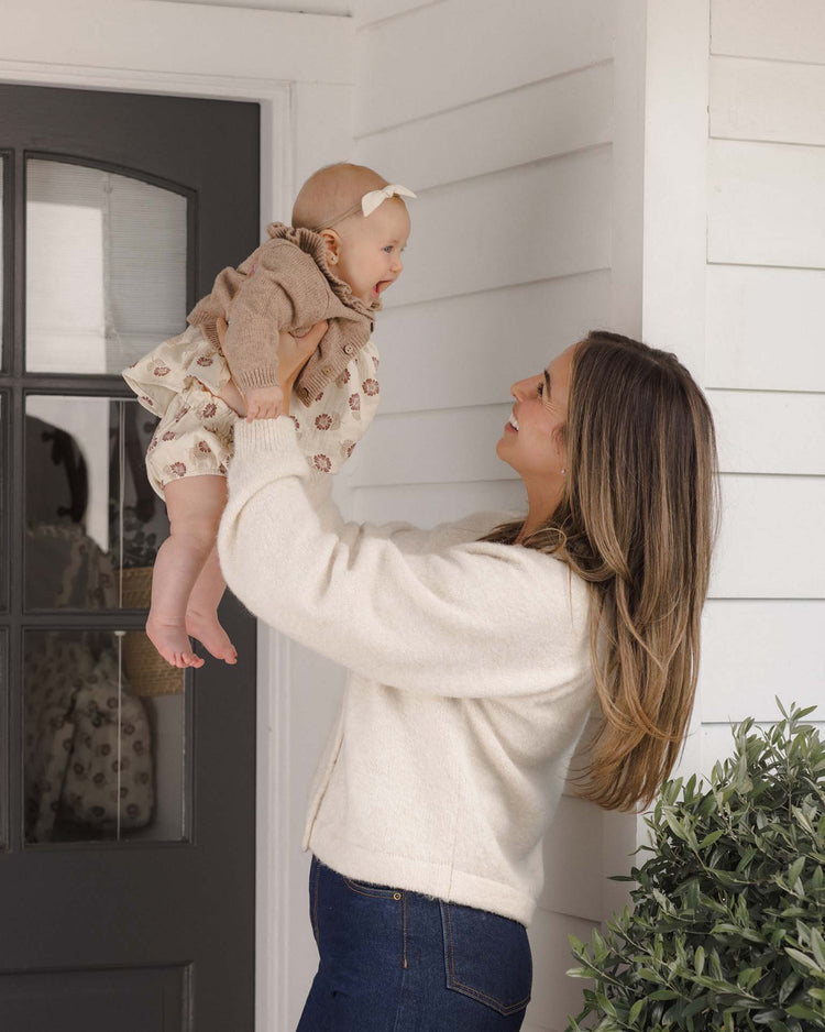 A woman with long hair, wearing the Quincy Mae ruffle collar cardigan in heathered almond and jeans, smiles as she lifts a baby dressed in a beige outfit and headband outside a white house with a glass door and nearby green plant.