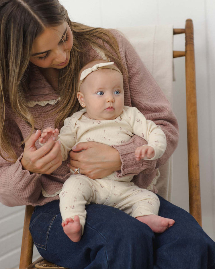 A woman sits on a wooden chair, holding a baby in her lap. The baby wears the quincy mae ribbed tee + legging set in fall ditsy with a matching headband, and both have relaxed expressions. The background is simple and neutral.