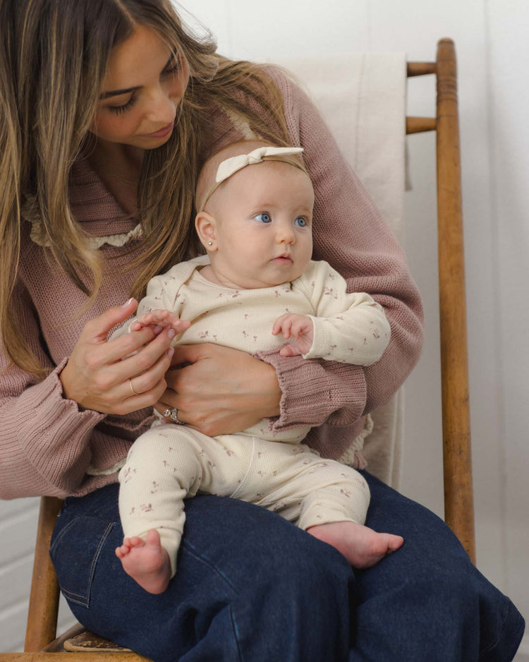 A woman wearing a mauve sweater and jeans holds a baby dressed in the quincy mae ribbed tee + legging set in fall ditsy, complete with a matching bow headband. They sit on a wooden chair, and the baby, cozy in the outfit, looks slightly to the side.