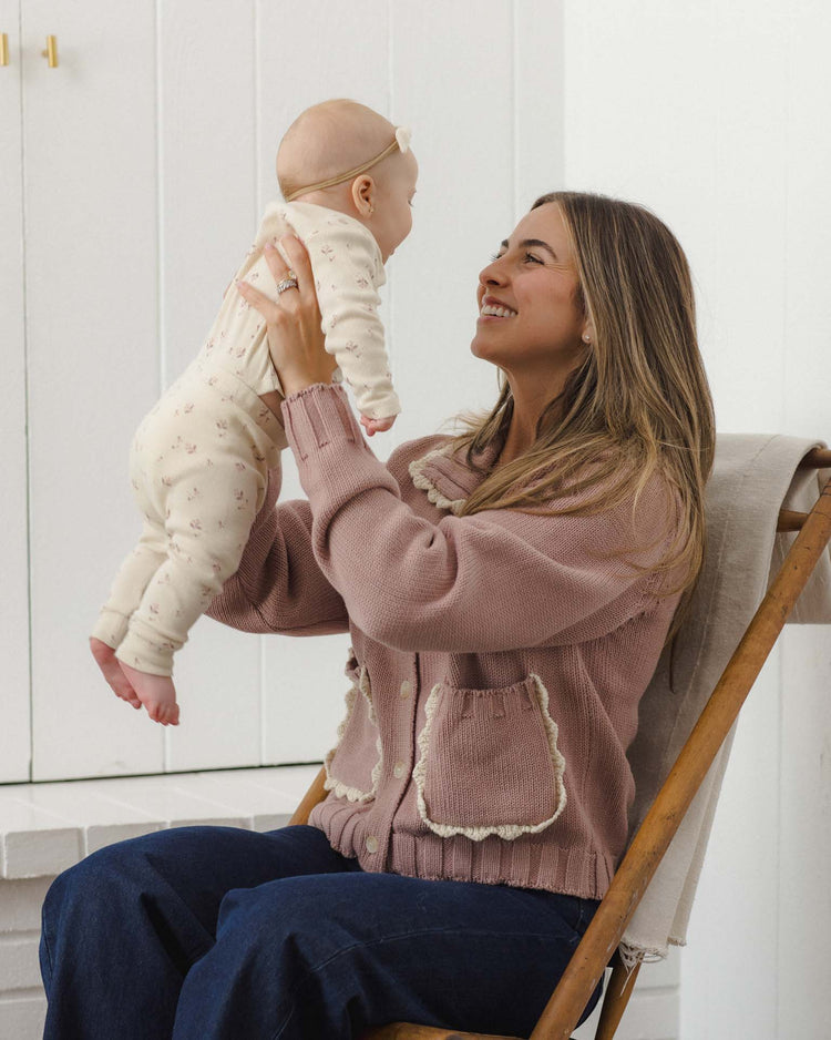 A woman seated on a wooden chair smiles as she lifts a happy baby facing her, dressed in the quincy mae ribbed tee + legging set in fall ditsy. The scene is bright and minimal.
