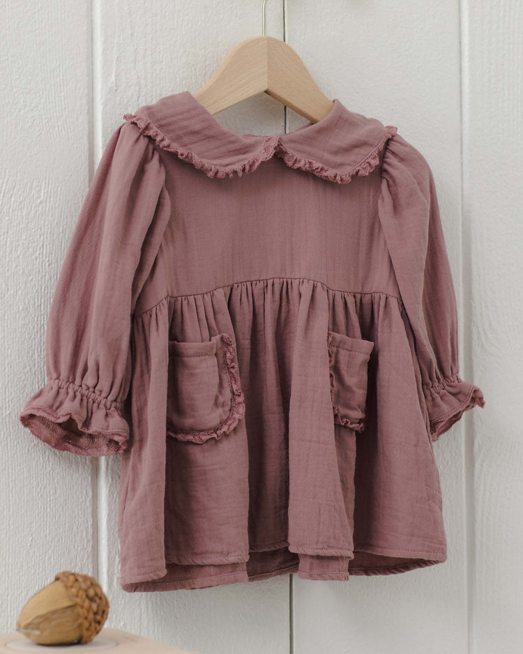The marisol dress in mulberry by quincy mae is a mauve long-sleeve organic cotton dress for toddlers, featuring a ruffled Peter Pan collar and cuffs, two front pockets, and a gathered waist. It is displayed on a wooden hanger against a white wall.