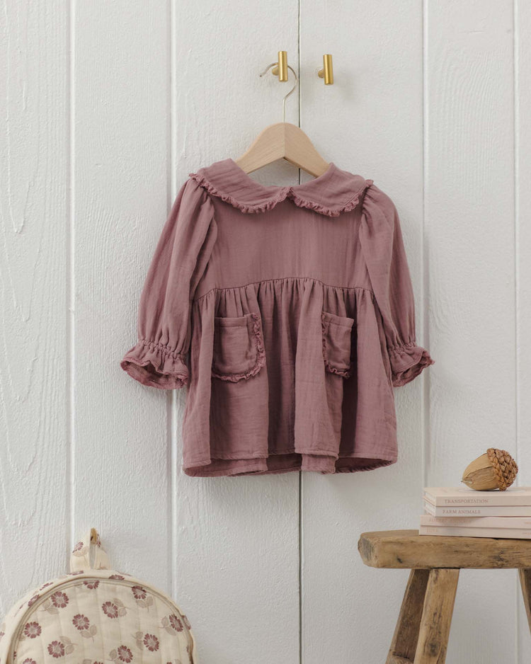 The marisol dress in mulberry by quincy mae—a long-sleeve organic cotton dress featuring a ruffled Peter Pan collar and convenient pockets—hangs on a wooden hanger against a white paneled wall. Nearby, a wooden stool holds books, and a floral-patterned backpack rests beside it.