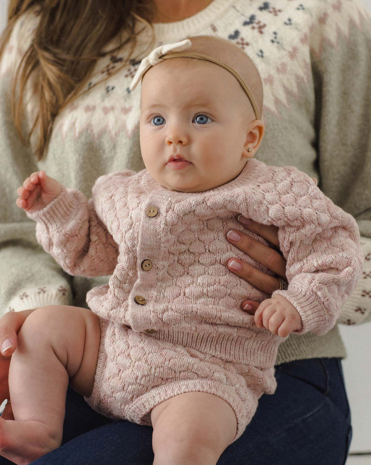A baby in a dusty rose knit outfit and headband sits on a woman’s lap. The woman wears a patterned sweater and blue jeans. Both are facing forward, and the baby has blue eyes and a curious expression. The quincy mae knit bloomer in dusty rose speckle is crafted from soft organic cotton, adding comfort to her adorable look.