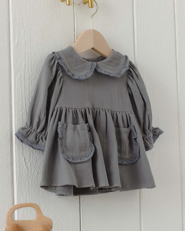 A marisol dress in slate by quincy mae, featuring long sleeves, ruffled cuffs, a scalloped Peter Pan collar, and two front pockets, crafted from soft organic cotton, is displayed on a wooden hanger against a white wall.
