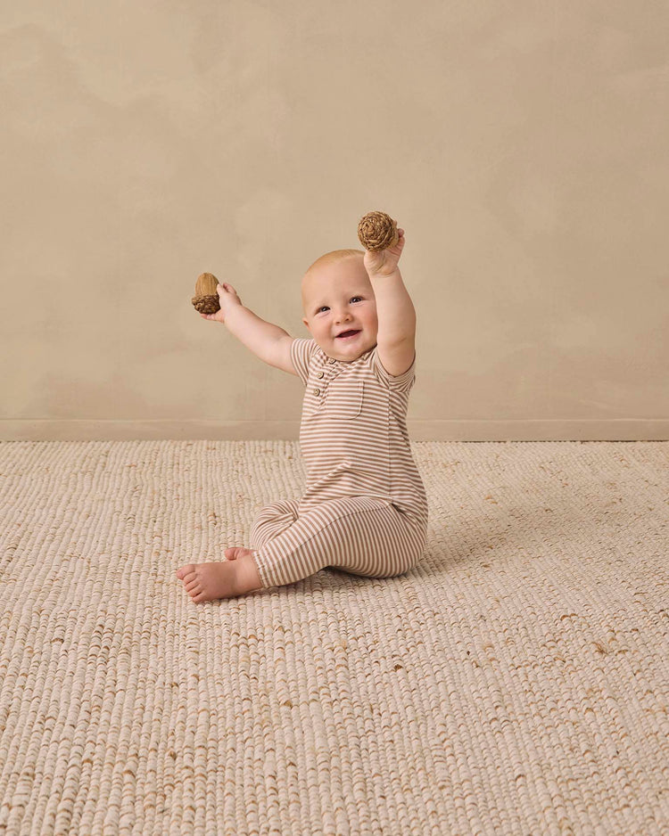 A smiling baby wearing the quincy mae short sleeve jumpsuit in almond stripe sits on a textured rug, holding two wooden toys with both hands raised.