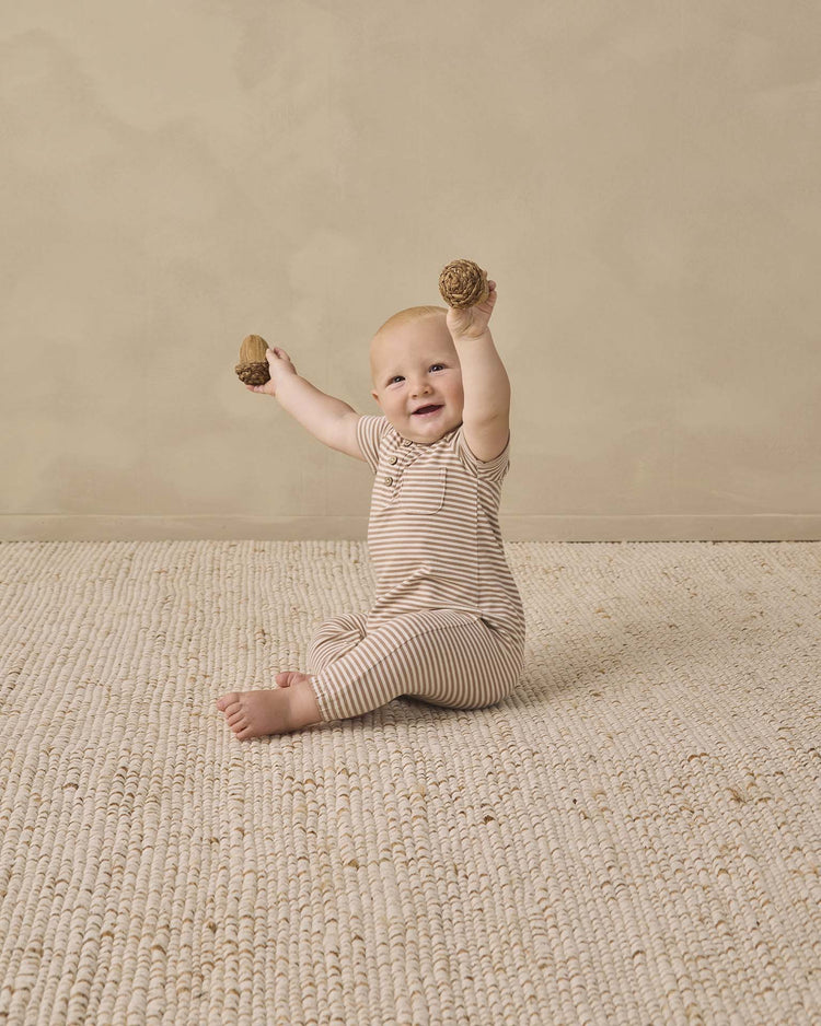 A smiling baby wearing the quincy mae short sleeve jumpsuit in almond stripe sits on a textured rug, holding two wooden toys with both hands raised.