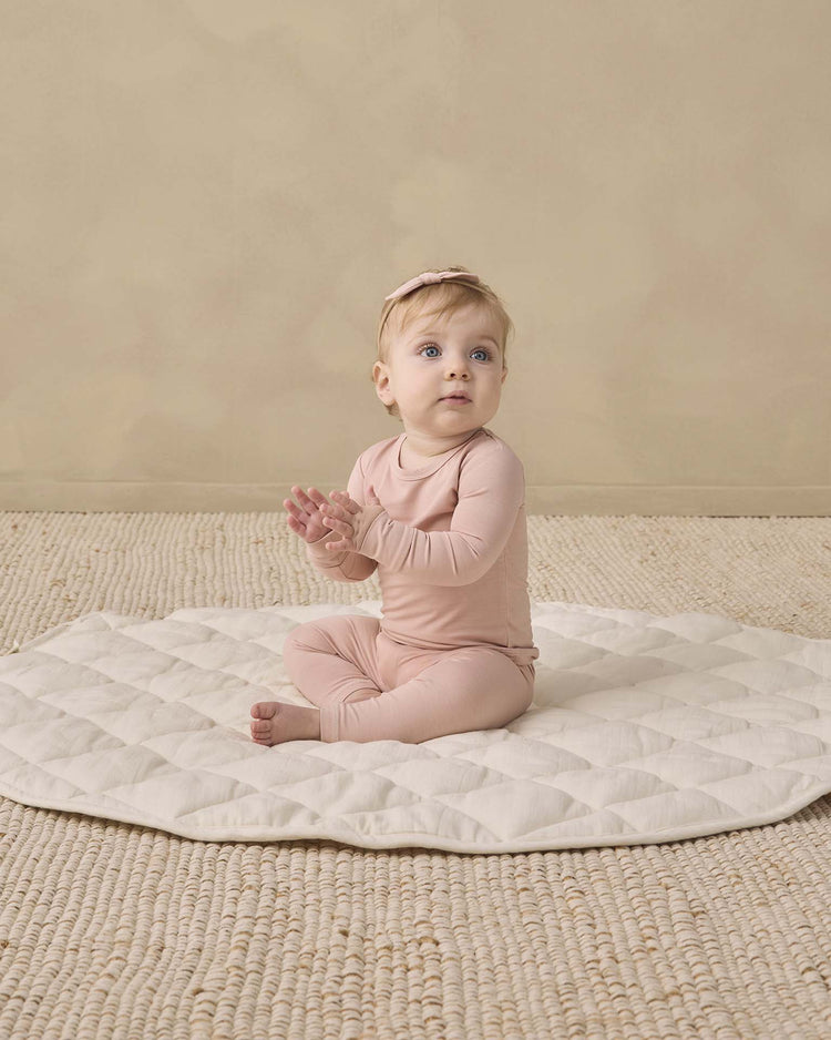 A baby wearing the quincy mae bamboo pajama set in dusty rose and a matching headband sits on a textured round mat, gazing slightly upward with hands together against a backdrop of neutral beige tones.