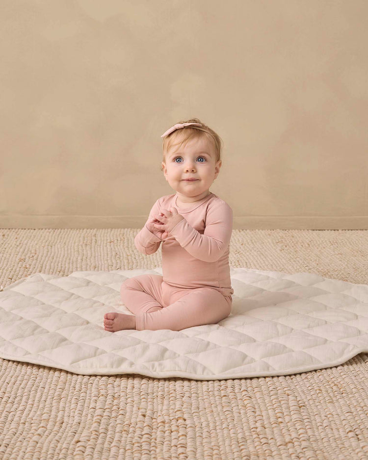 A baby wearing the quincy mae bamboo pajama set in dusty rose, paired with a matching headband, sits on a quilted cream mat with hands clasped, looking up. The background features a beige textured rug.
